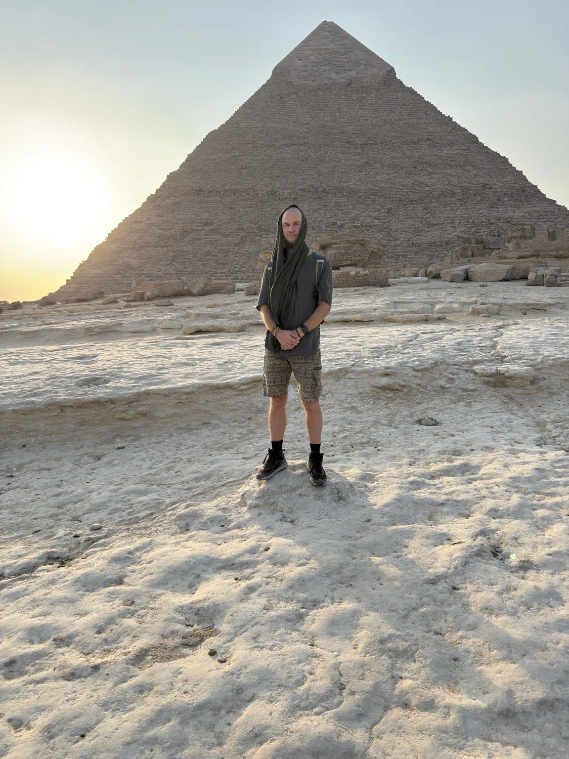 Tyler standing hooded on pale bedrock, the pyramid of Khafre filling the frame behind him, sun low at left