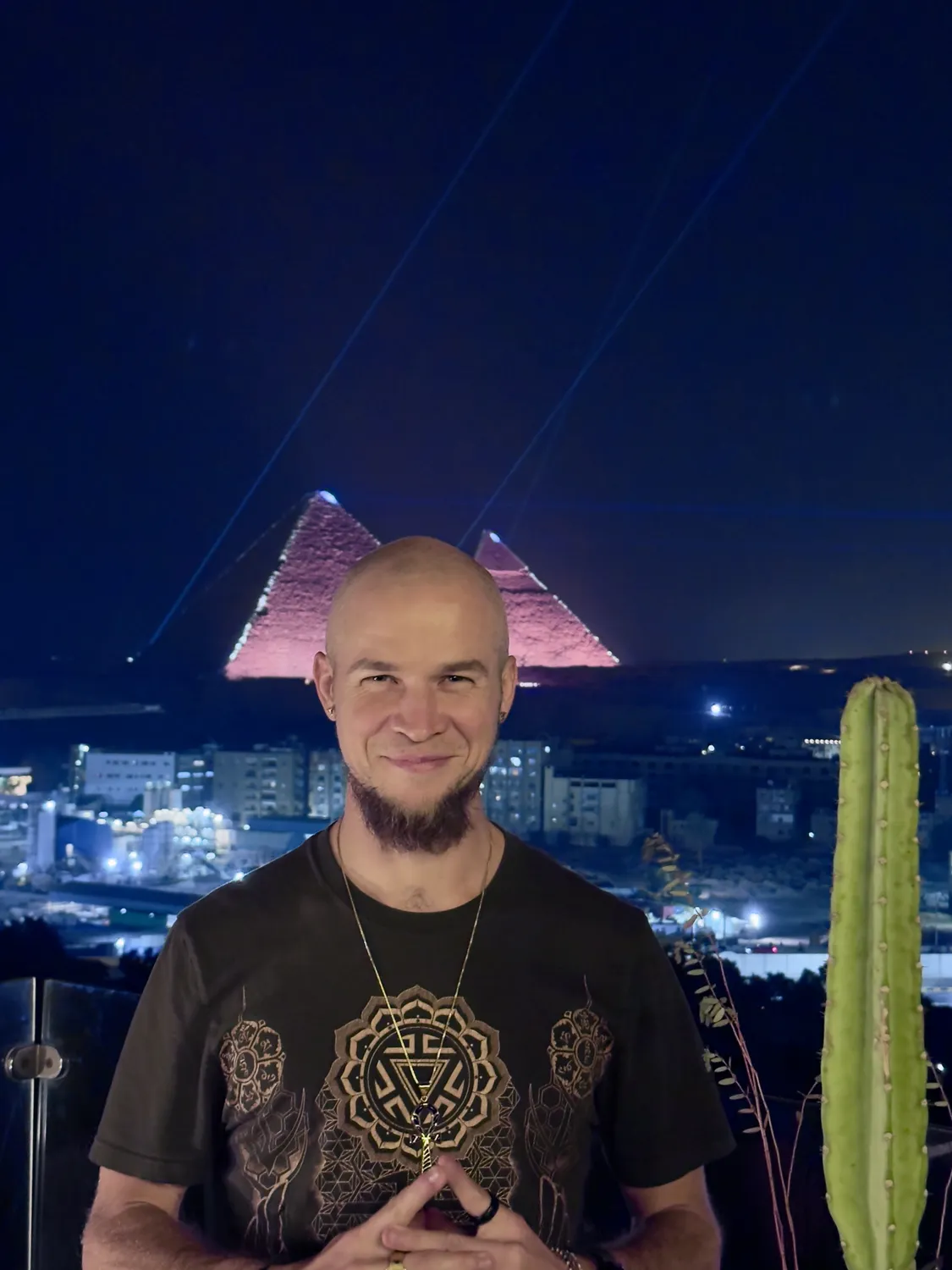 Tyler smiling with hands together at his chest, pink-lit pyramids and laser beams in the night sky behind him