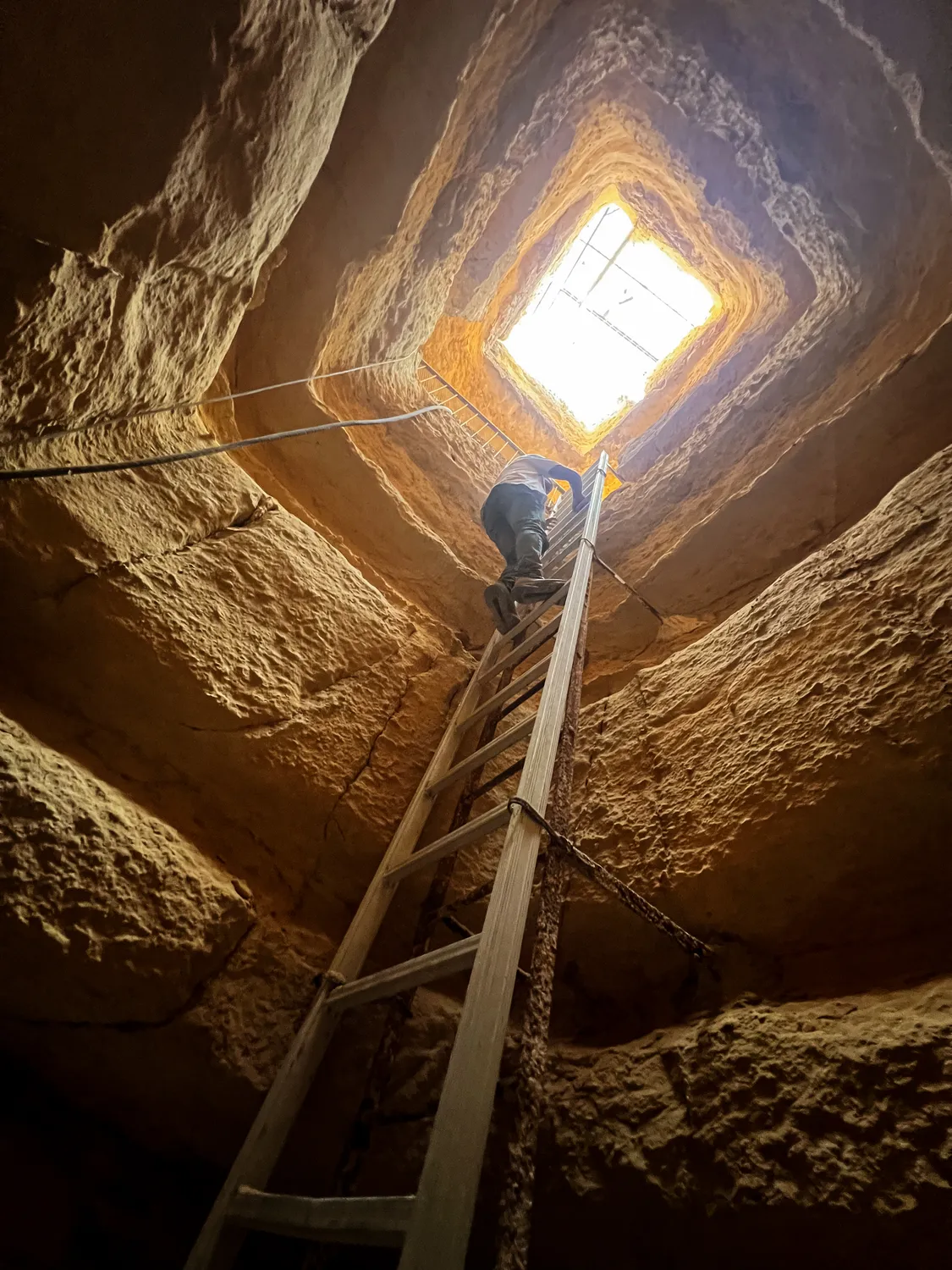 Looking up a rock shaft as a climber on a ladder rises toward a square of daylight