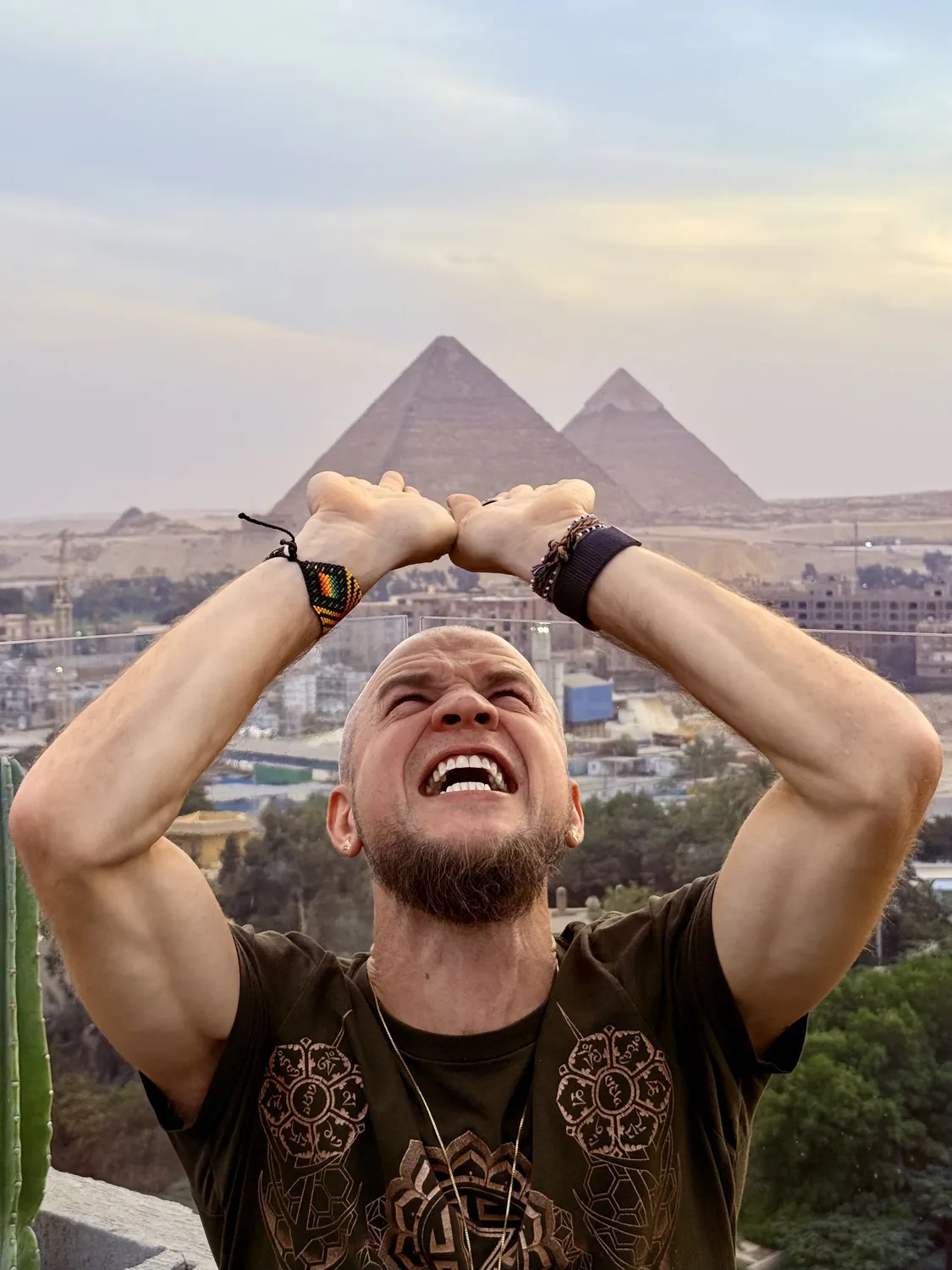 Tyler on a rooftop with his arms raised in a triangle over his head, beaming, two Giza pyramids behind him