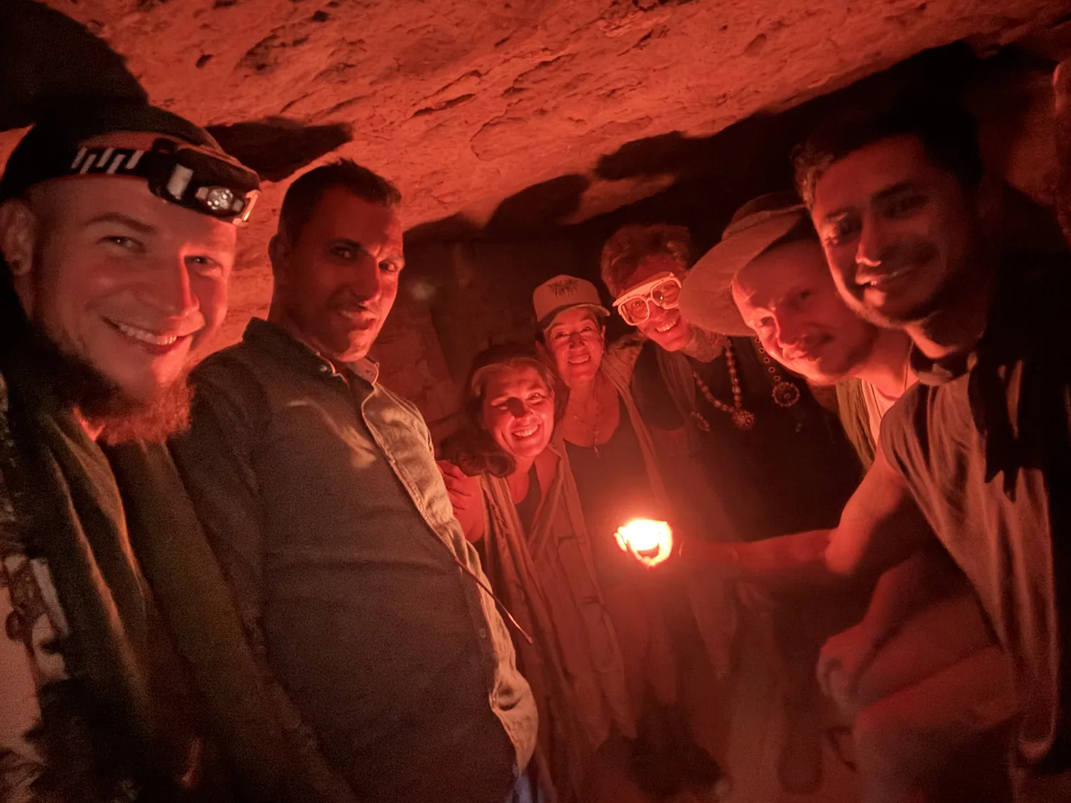 A group lying on a chamber floor in red light, faces turned up toward the camera, a small flame at centre