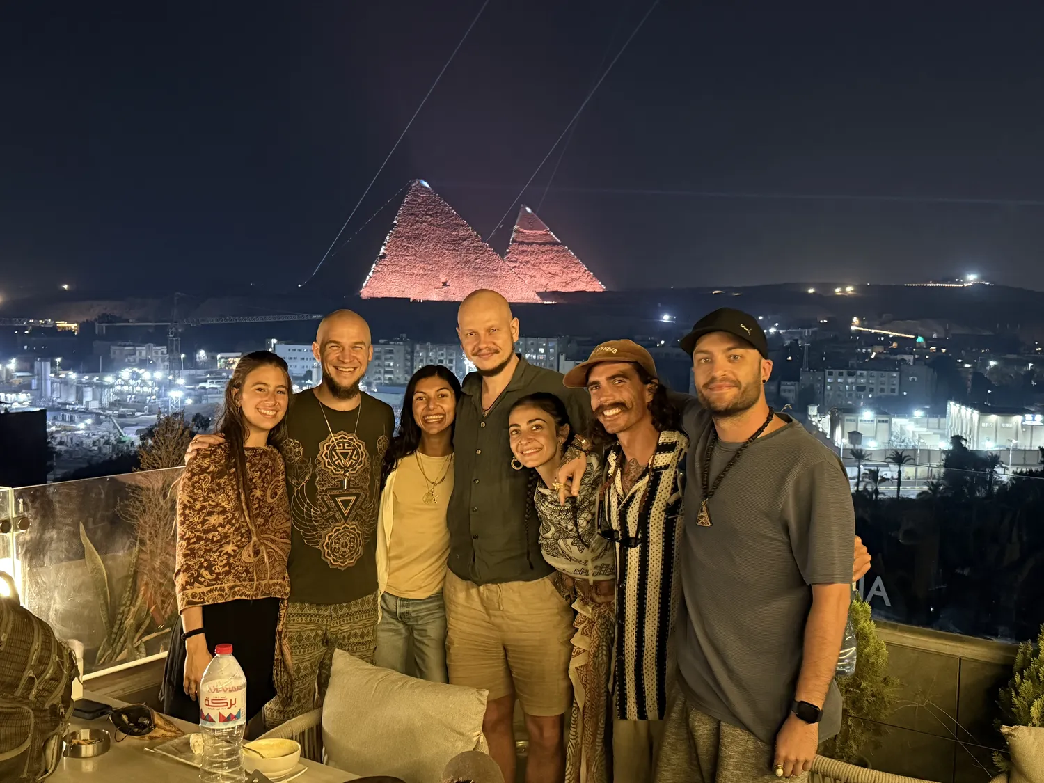 Seven people standing together on a rooftop at night, pink-lit pyramids behind them