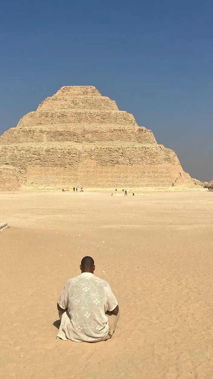 Tyler seen from behind, seated cross-legged on the sand facing the Step Pyramid of Djoser
